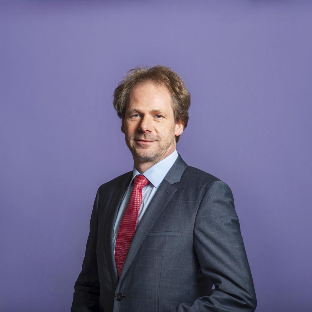 Foto van Bart Snels, een witte man. Hij draagt een zwart pak met een rode stropdas, heeft donkerblond haar en een vriendelijke uitdrukking op zijn gezicht.