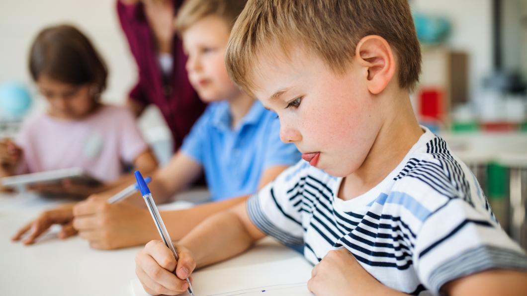 Een groep kleine kinderen in de klas die leren schrijven, met een jongetje met geconcentreerde blik vooraan in beeld