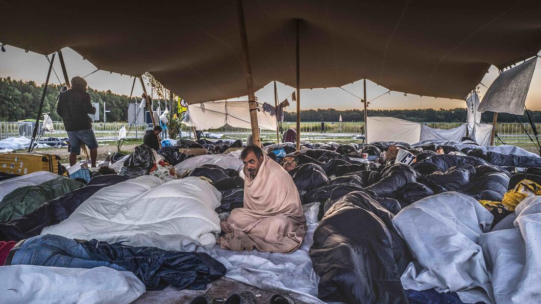 Een heleboel mensen liggen dicht op elkaar onder een zeil te slapen. Ze zijn vluchtelingen die liggen te wachten voor de poort van Ter Apel. In het midden zit een man, hij is gewikkeld in een deken en kijkt voor zich uit. 