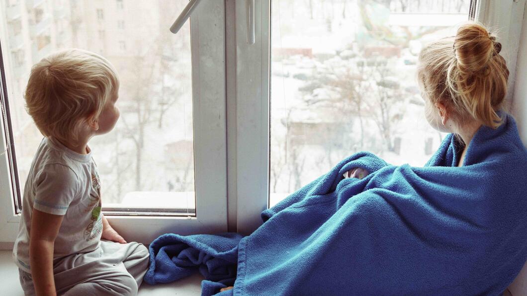 Twee kleine kinderen zitten voor een raam. Buiten is het koud. Het rechterkind heeft een blauwe deken om zich heen geslagen