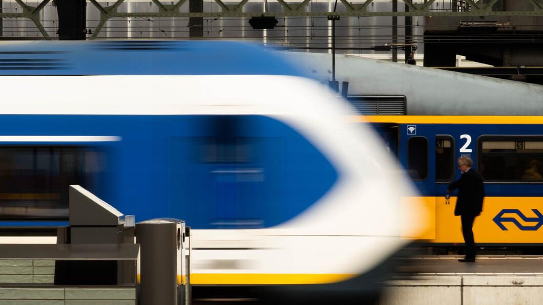 Beeld van een station, waar een sprinter voorbij rijdt terwijl een intercity trein stilstaat.