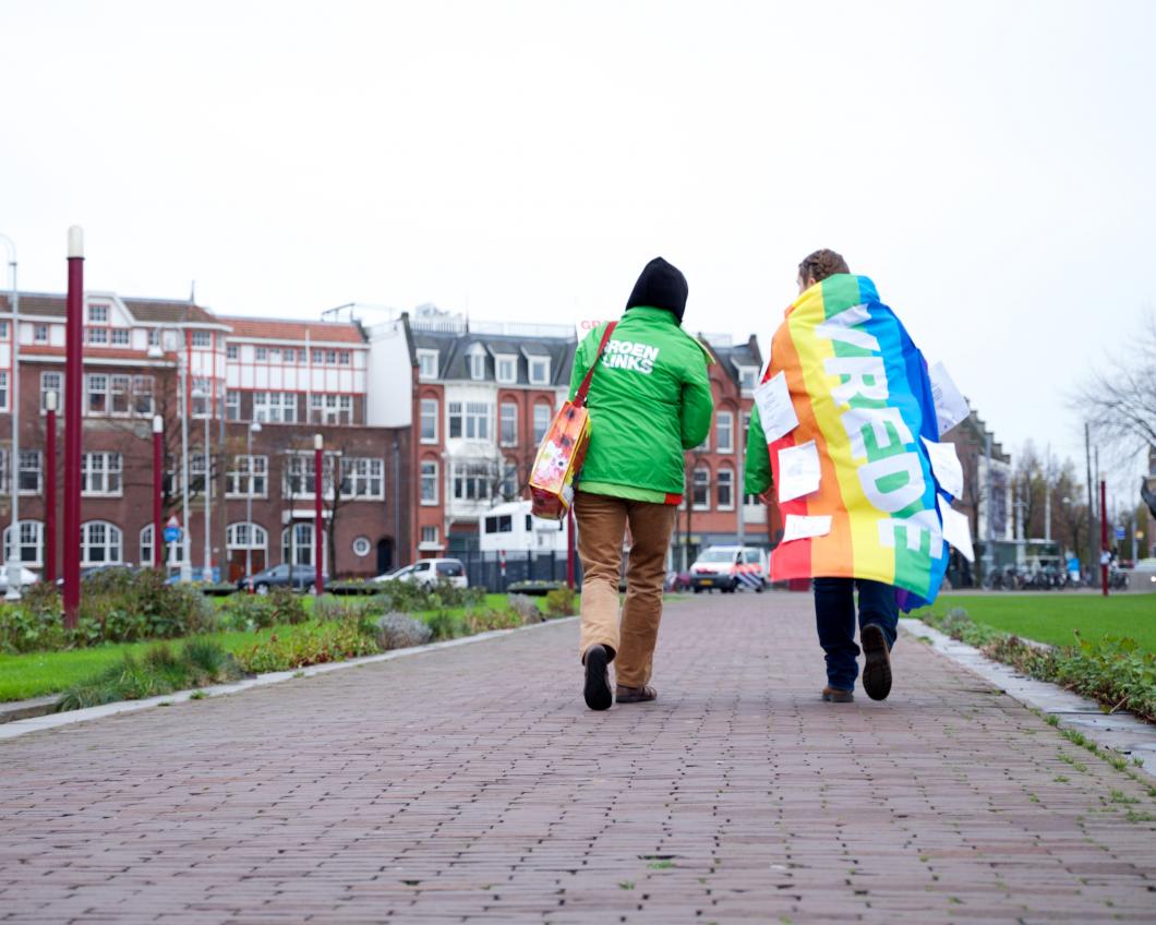 Twee vrijwilligers lopen over straat, een heeft de regenboogvlag met het woord 'vrede' over de schouders geslagen.