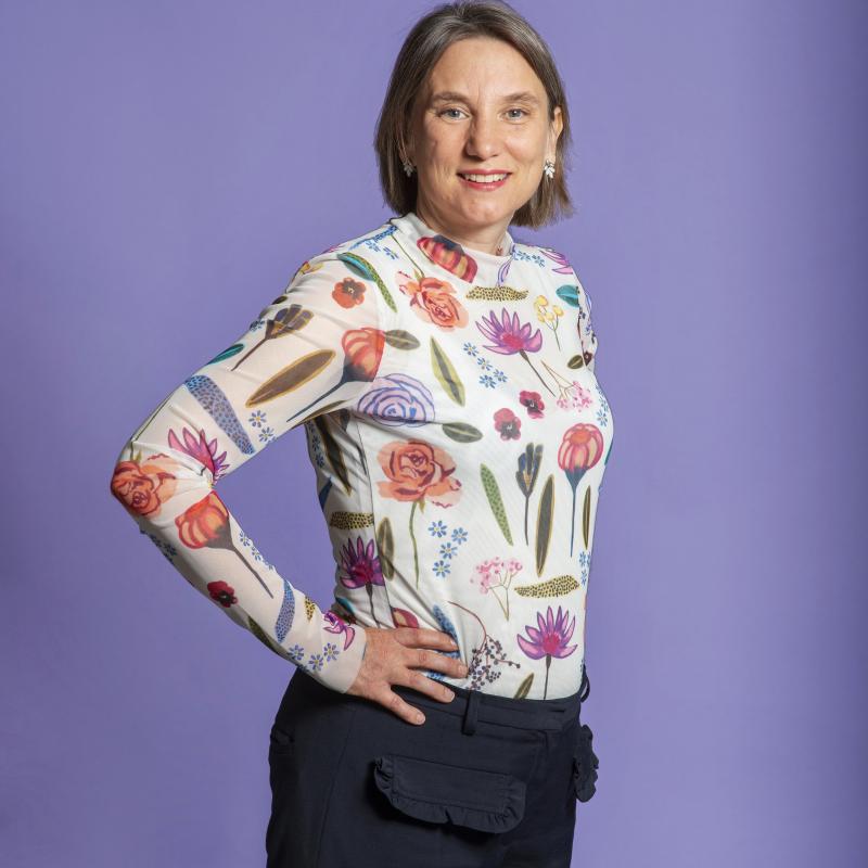 Foto van Laura Bromet, een witte vrouw. Ze draagt een wit shirt met bloemetjes in verschillende kleuren en draagt een zwarte broek, heeft donkerblond haar en lacht.